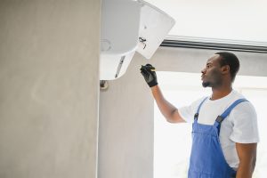 A professional HVAC technician in blue overalls using a screwdriver to service and repair a wall-mounted ductless mini-split air conditioning unit indoors.