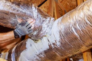 A close-up of a worker wearing a grey protective glove connecting and insulating silver flexible air ductwork onto a main ventilation plenum in a home attic space.
