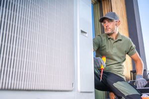 A professional HVAC technician in safety glasses and work gloves holding a screwdriver while performing a maintenance inspection on a residential outdoor air conditioner condenser and heat pump unit.