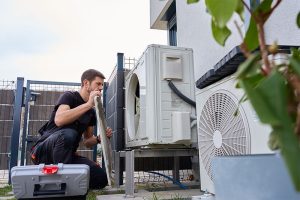 A professional HVAC technician servicing a residential central air conditioning condenser unit outside a home, prepared with a gray toolbox to inspect and repair the AC system.
