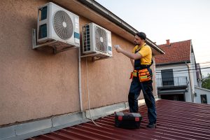 A professional HVAC technician standing on a red corrugated roof to perform a thermal diagnostic check on wall-mounted outdoor air conditioning condenser units using an infrared thermometer.
